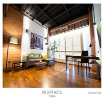 An executive suite at The Loft Hotel in Yangon with a brick wall, a large window, a sofa, a coffee table, a desk, and a potted plant.