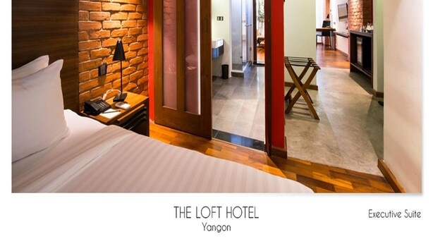 Una suite ejecutiva en The Loft Hotel, Yanjon, con una pared de acento de ladrillo, una cama, un escritorio con lámpara y vistas al interior de la habitación.