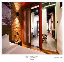 An executive suite at The Loft Hotel, Yanjon with a brick accent wall, a bed, a desk with a lamp, and a view into the room.