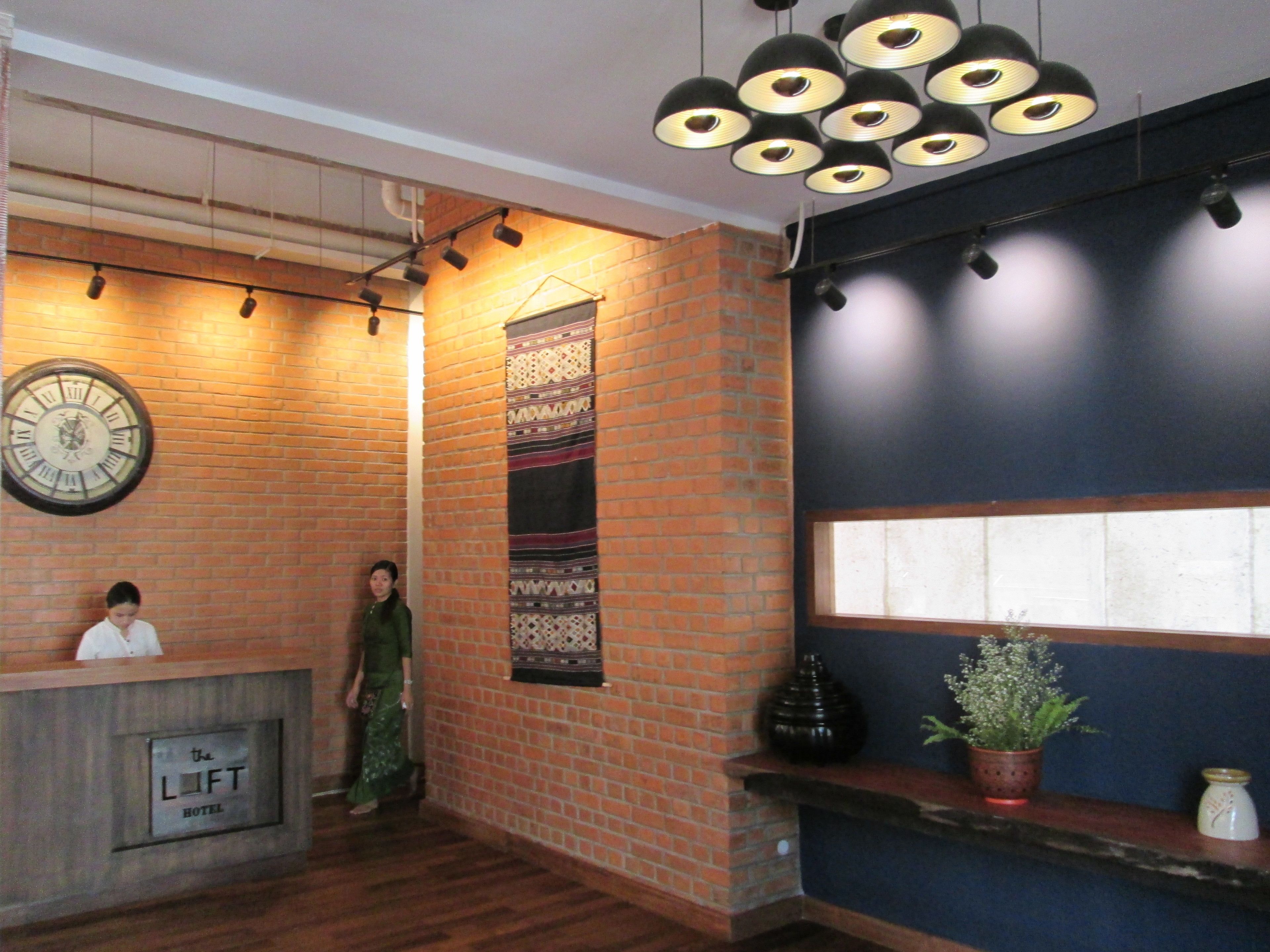A hotel reception area with a brick wall, a large clock, a patterned tapestry, and a reception desk labeled "The Loft Hotel".