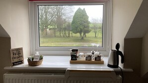 Un coin cuisine avec une fenêtre donnant sur un champ verdoyant et des arbres. Sur le rebord de la fenêtre, on trouve divers objets, notamment un panier, une pancarte indiquant « Le mot de passe du Wi-Fi » et une machine à café.