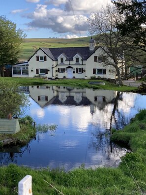 A two-story white building with a symmetrical facade, a central entrance, and a reflection in a calm water body in front.