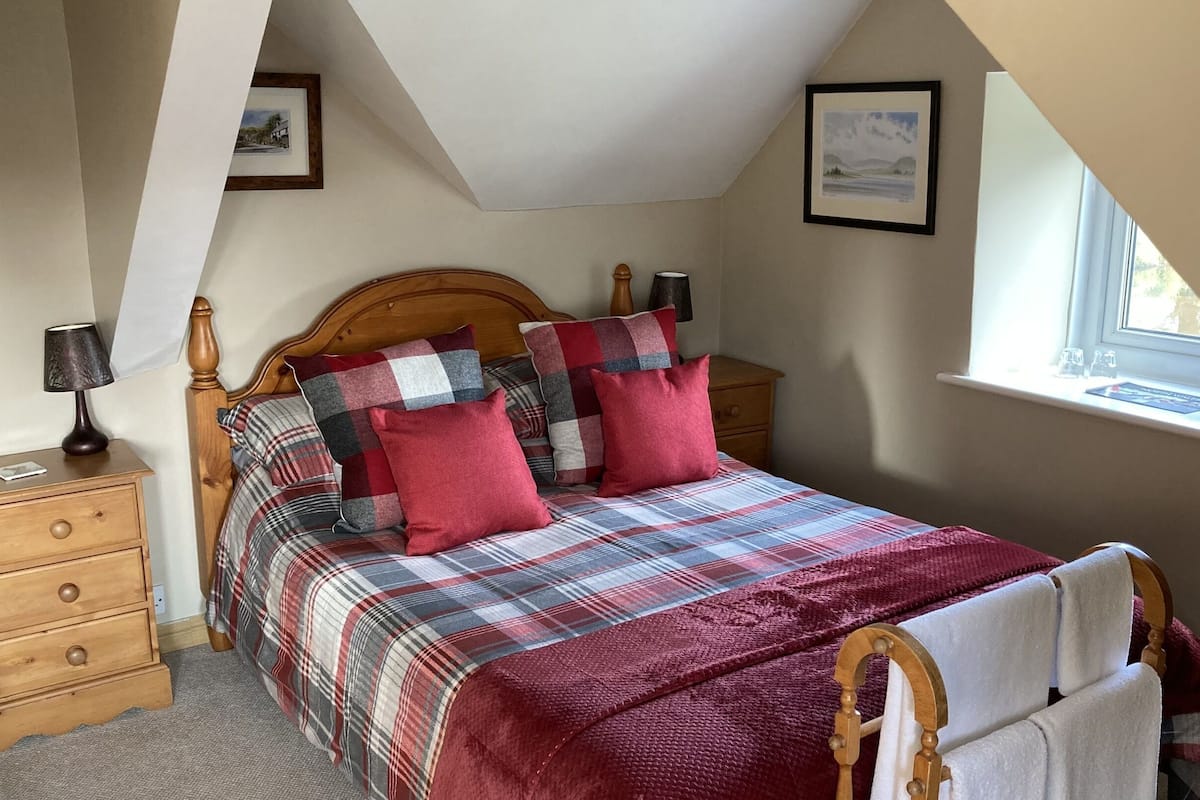 Une chambre à coucher avec un lit en bois, une literie à motifs écossais, une table de chevet, une lampe et une fenêtre donnant sur un beau paysage.