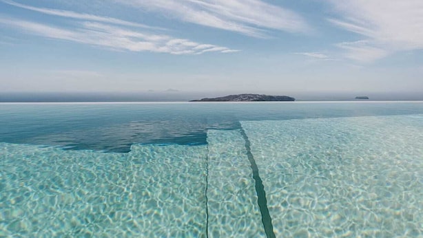 On aperçoit clairement une plage avec une jetée en bois qui s’avance dans l’eau, une île au loin et une mer calme sous un ciel partiellement nuageux.