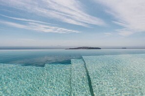 On aperçoit clairement une plage avec une jetée en bois qui s’avance dans l’eau, une île au loin et une mer calme sous un ciel partiellement nuageux.