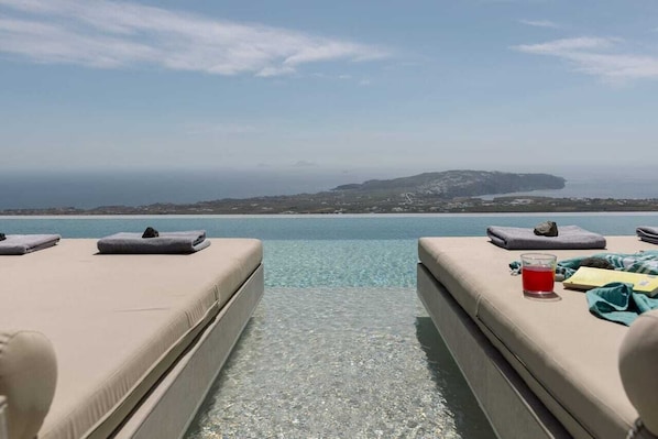 A clear view of a poolside area with lounge chairs and a glass of red drink, overlooking a coastal landscape.