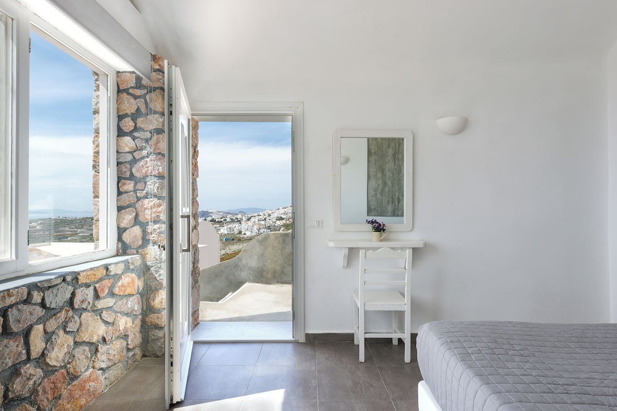 A bedroom with a stone wall, a white dresser, a bed, and a view of a town through an open door.