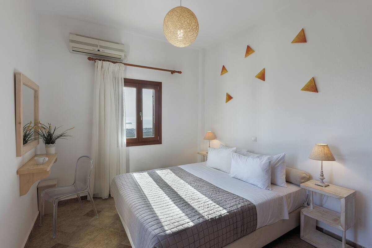 A bedroom with a bed, bedside lamps, a chair, a desk, a potted plant, and a window with curtains.