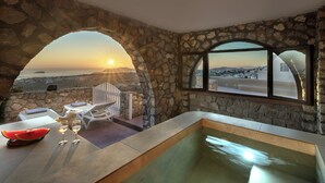 Une pièce aux murs de pierre offrant une vue sur le coucher de soleil sur la mer, avec une piscine et une table sur laquelle se trouvent une pastèque et des verres.
