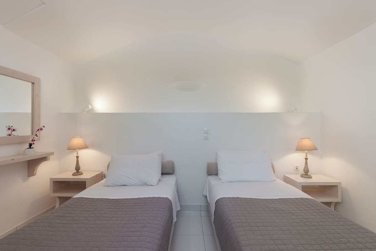 A hotel room with two beds, each with a bedside table and lamp, a small shelf with flowers, and a mirror on the wall.