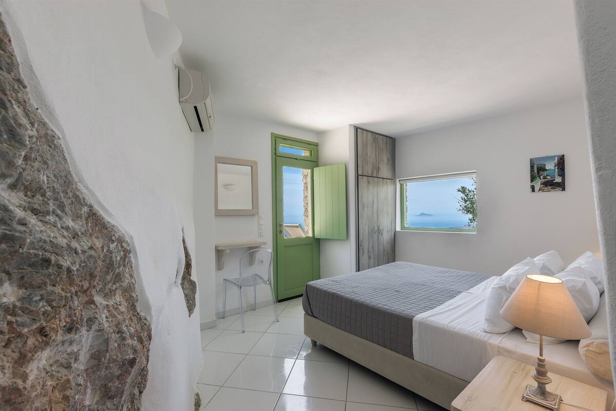 A bedroom with a bed, a bedside lamp, a chair, a green door, and a window with a view.