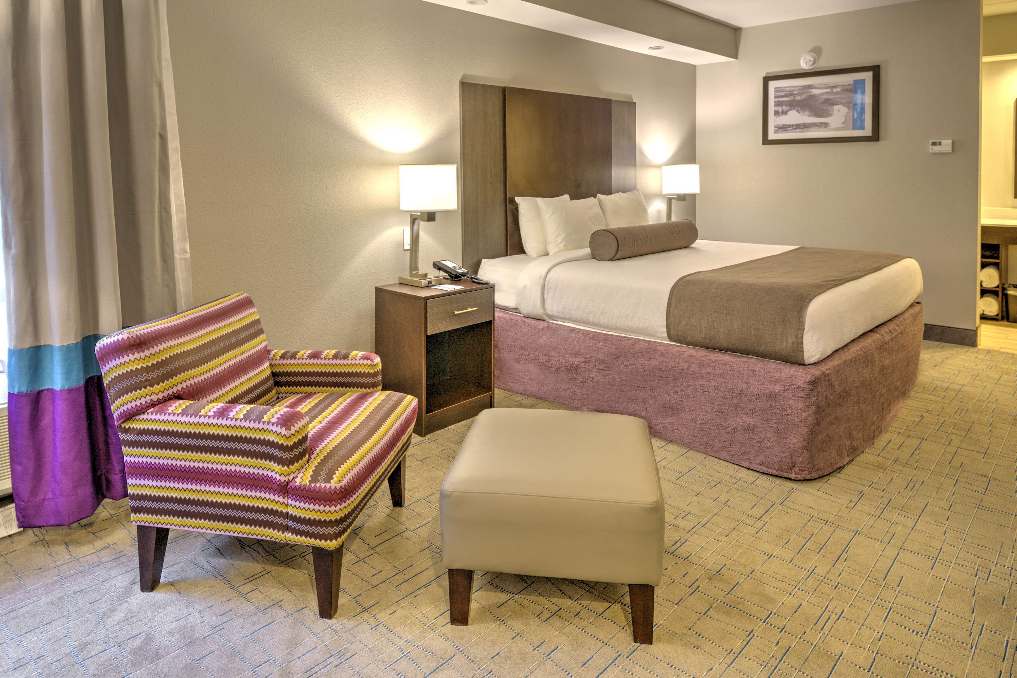 A hotel room with a bed, a chair, an ottoman, a nightstand, and a lamp.