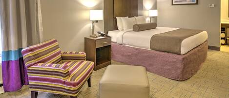 Una habitación de hotel con cama, sillón, reposapiés, mesita de noche y lámpara.