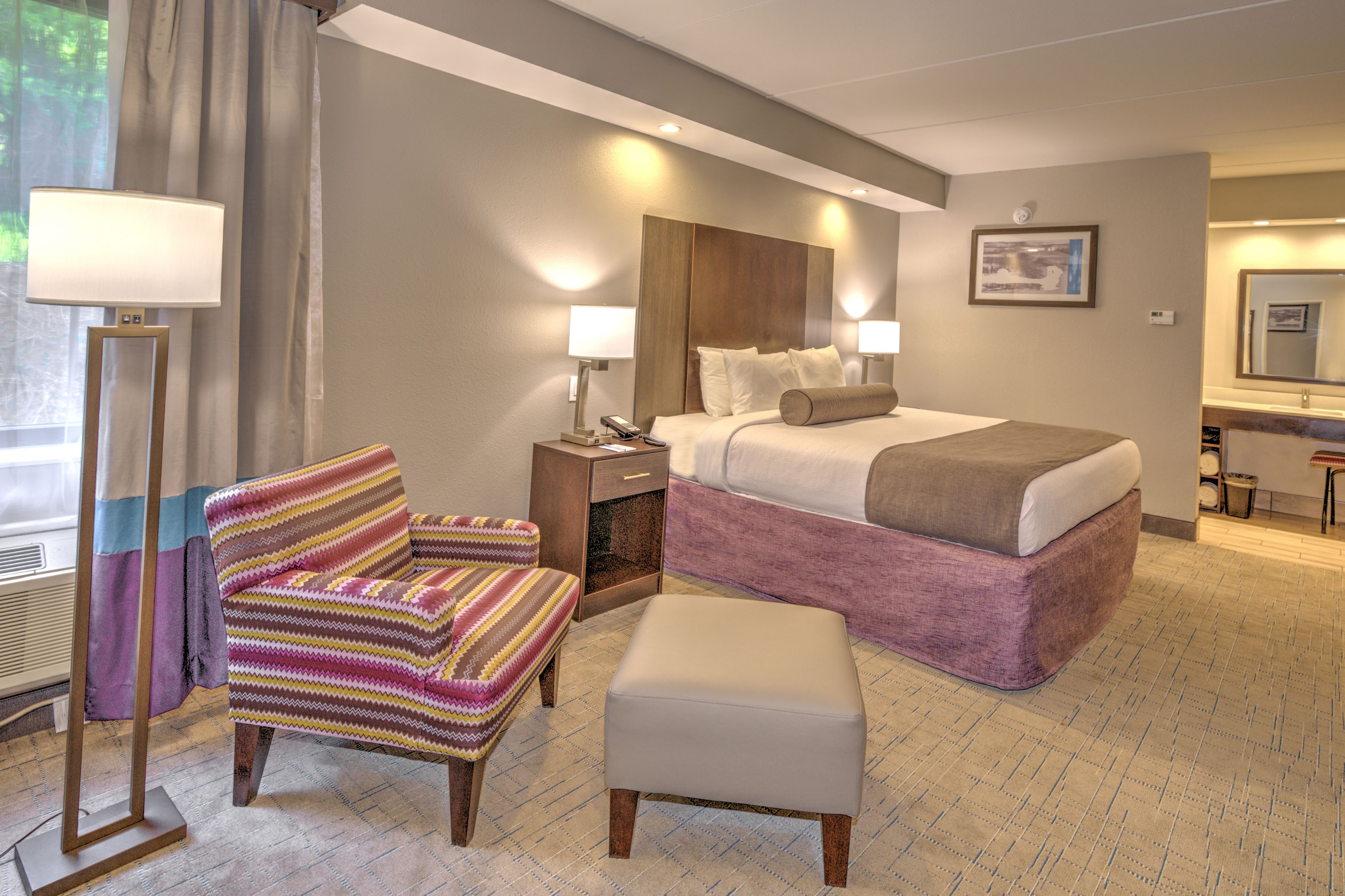 Ein Hotelzimmer mit Bett, Stuhl, Nachttisch mit Lampe und einem kleinen Tisch mit Spiegel.
