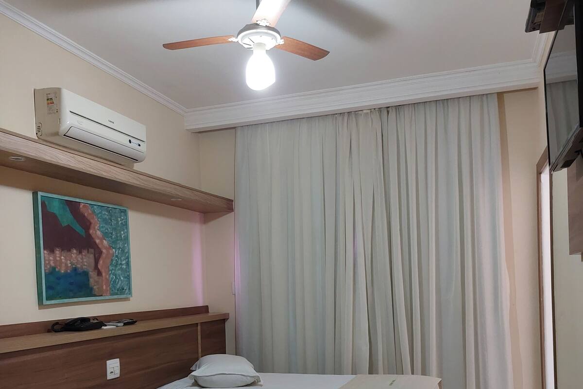 Habitación de hotel con dos camas, ventilador de techo, aire acondicionado, un cuadro y un ventanal grande con cortinas.