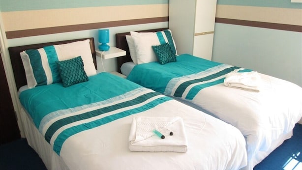 A hotel room with two beds, each with teal and white bedding, a small bedside table with a blue lamp, and a white towel on the bed.
