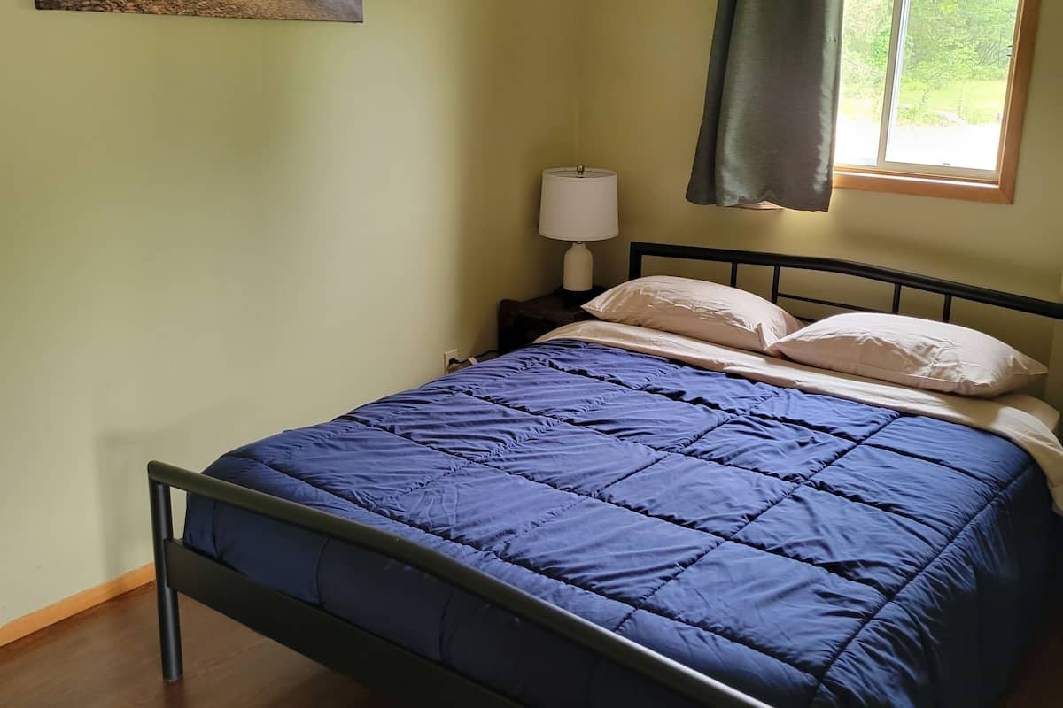 Un dormitorio con cama, mesita de noche, lámpara, ventana con cortinas y un cuadro en la pared.