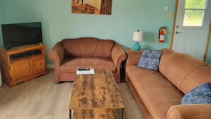 A living room with a television on a wooden stand, two brown sofas, a wooden coffee table, and a painting of a lakeside scene on the wall. - All Star Resort (South Algonquin)