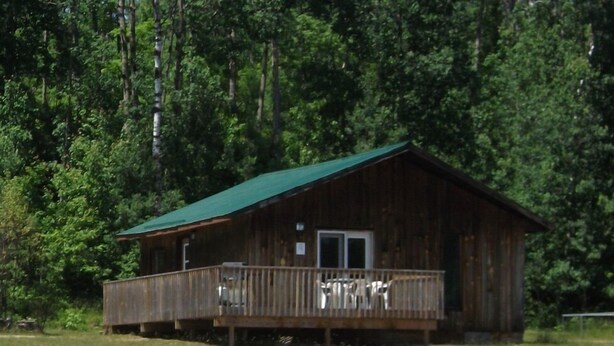 A wooden cabin with a green roof, a porch, and a small dock extending into a body of water. - All Star Resort (South Algonquin)