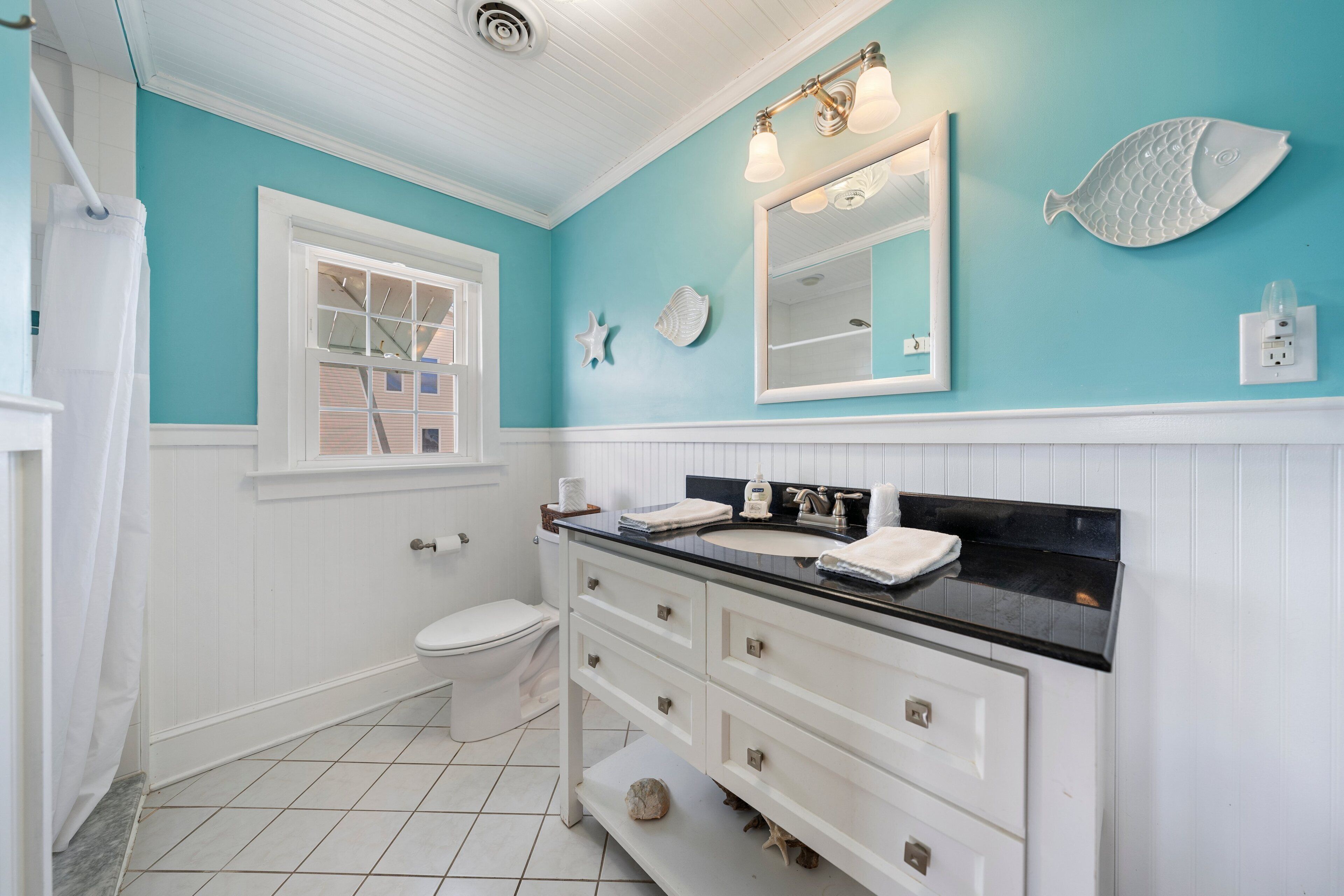 A bathroom with a white vanity, a sink, and a toilet. There are decorative shells on the wall and a window above the toilet.