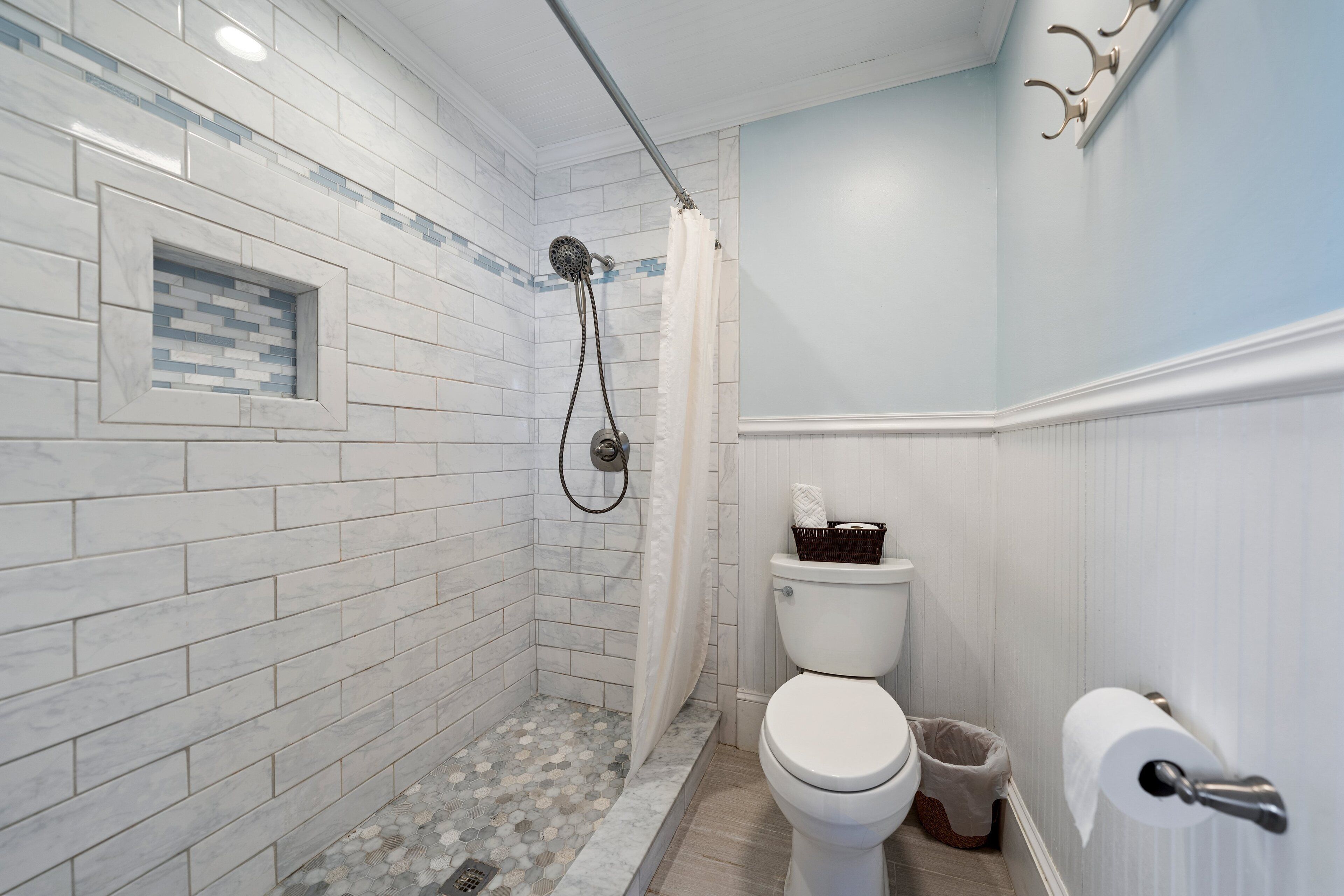 A bathroom with white tiled walls, a shower with a glass door, a toilet with a trash bin, and a towel rack.