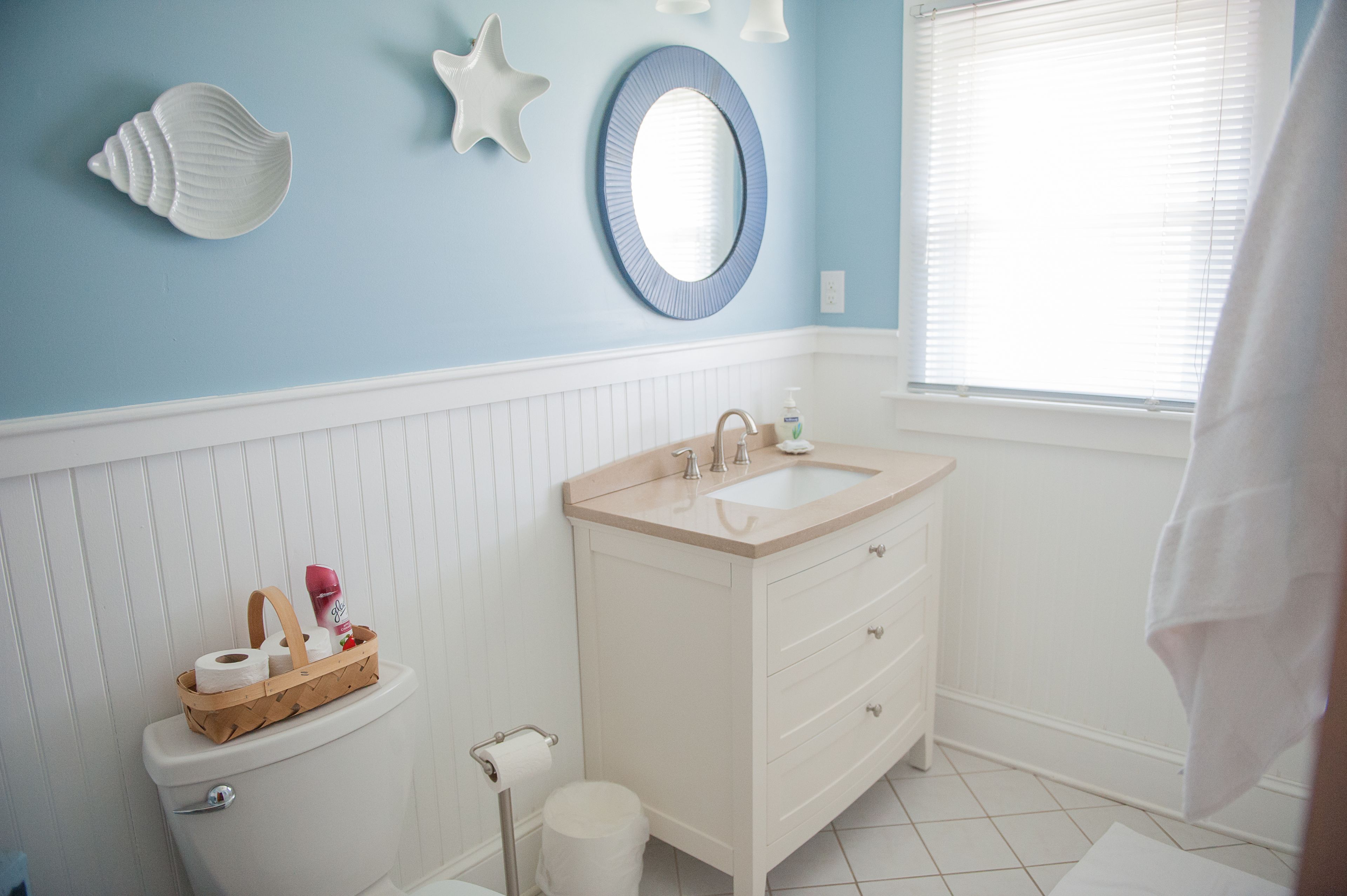 Un baño con inodoro, lavamanos y espejo. Hay decoraciones con conchas marinas en la pared y una ventana con persianas.