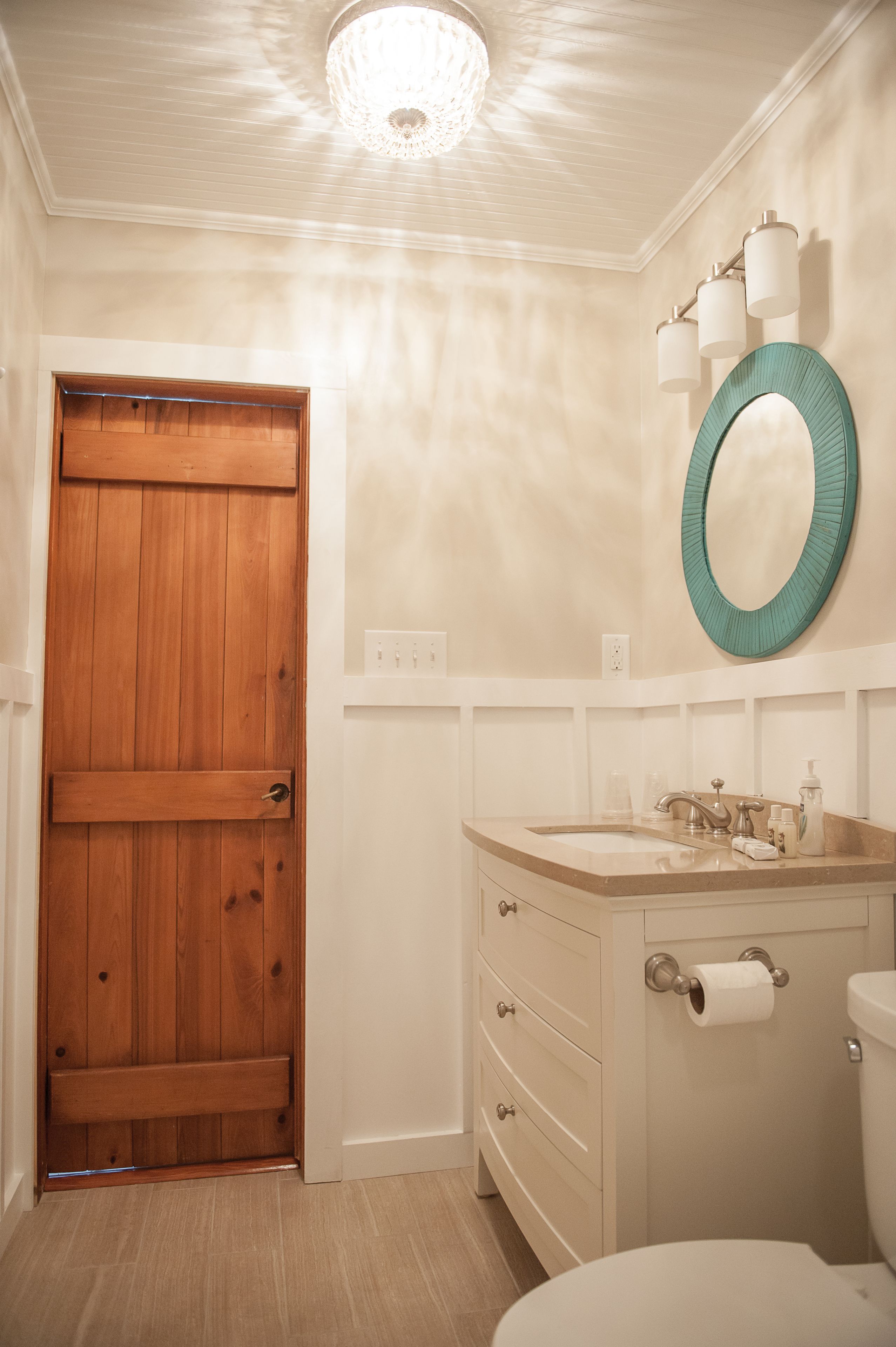 Um banheiro com porta de madeira, toucador branco com espelho e vaso sanitário.