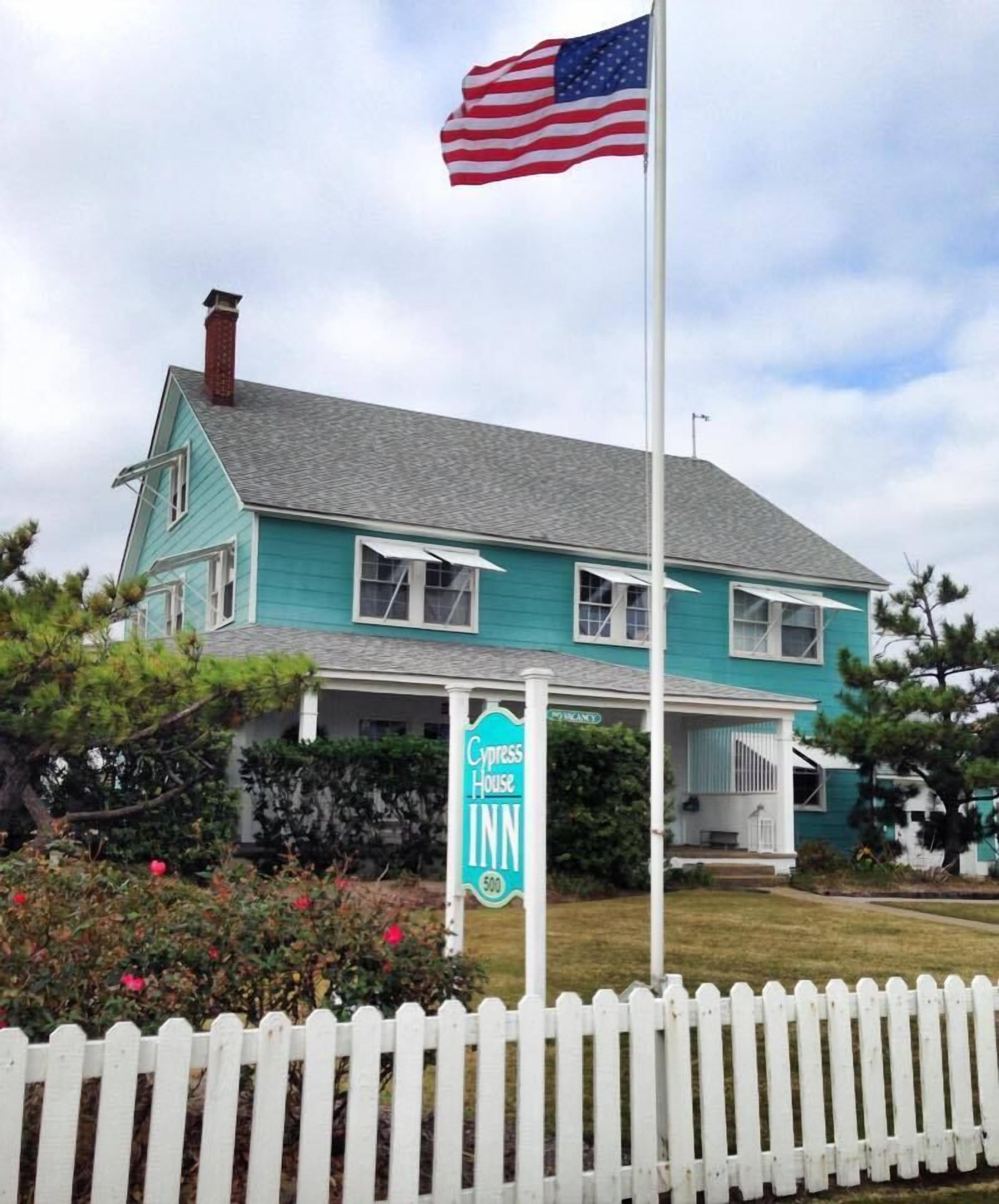 A turquoise two-story house with a white picket fence, an American flag, and a sign for Cypress House Inn.