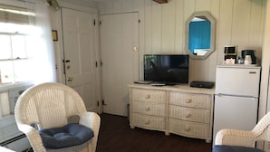 A room with a white wicker chair, a television on a dresser, and a refrigerator. - Spring Garden Inn (East Sandwich)