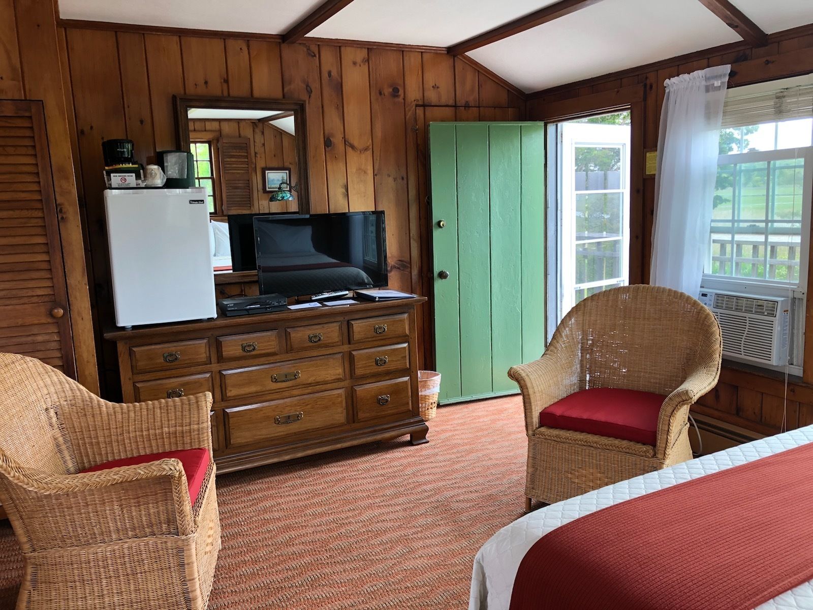 Um quarto de cabana aconchegante, com interior de madeira, uma porta verde, uma televisão em uma cômoda, cadeiras de vime e uma cama com um cobertor vermelho.