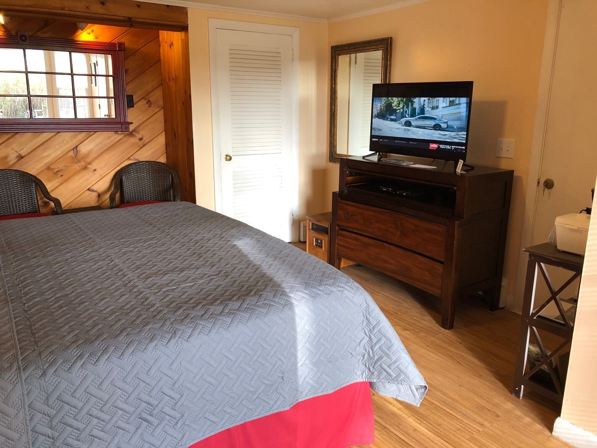 Een slaapkamer met een bed, een televisie op een houten dressoir, een deur en een raam met jaloezieën.