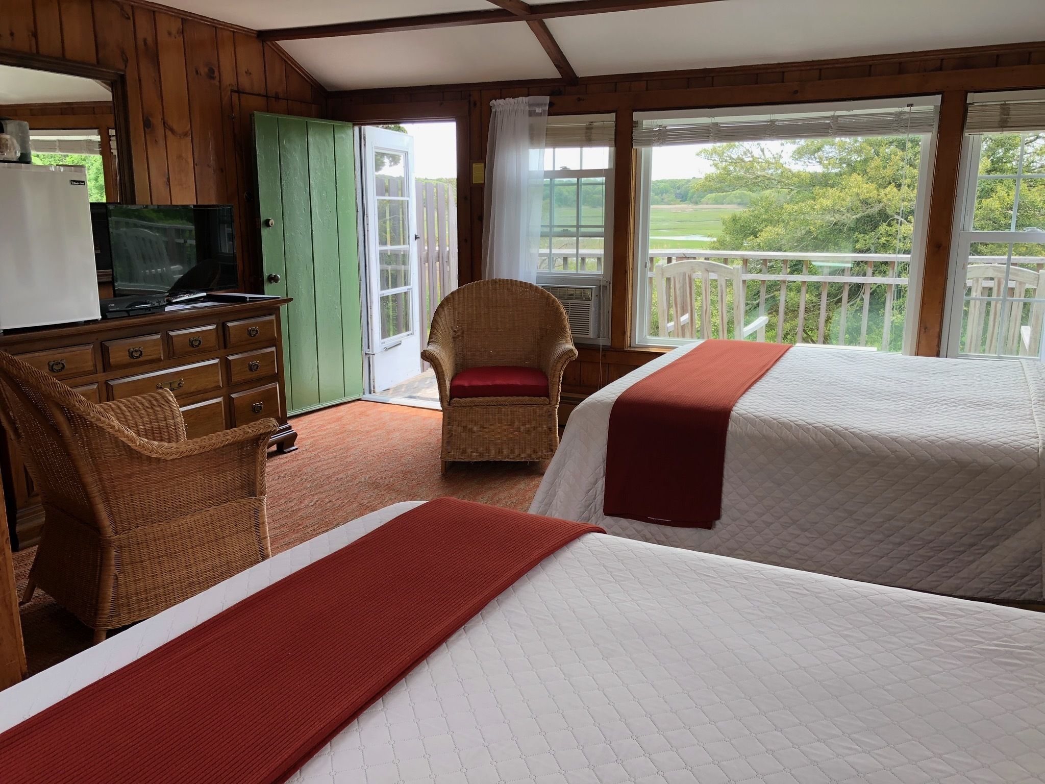 A room with two beds, a television, a wicker chair, and a balcony with a view of greenery.
