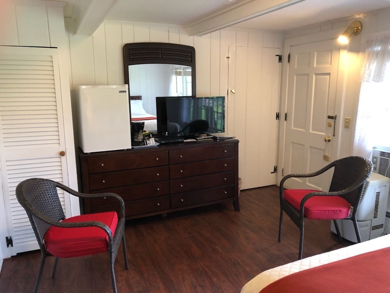 A room with a dresser, two wicker chairs with red cushions, a television, and a white door.