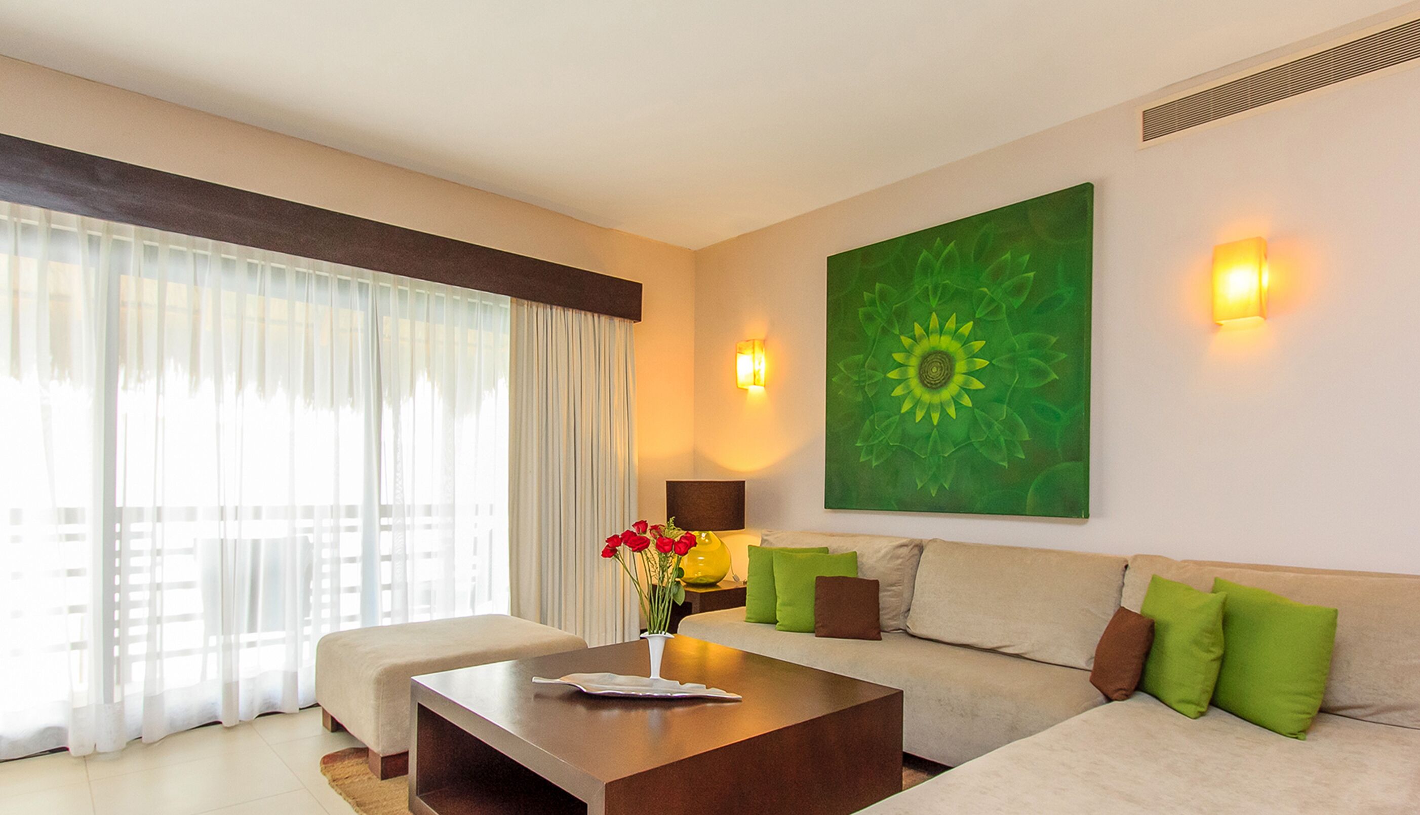 A modern hotel room with a sofa, a coffee table, a vase with flowers, and a large green floral painting on the wall.