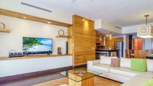 Una sala de estar moderna con un televisor de pantalla plana, una mesa de centro de vidrio, un sofá blanco y una cocina con armarios de madera y electrodomésticos de acero inoxidable.