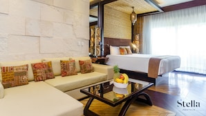 Habitación de hotel moderna con una cama grande, un sofá con cojines estampados, una mesa de centro de cristal con fruta y vistas al exterior.