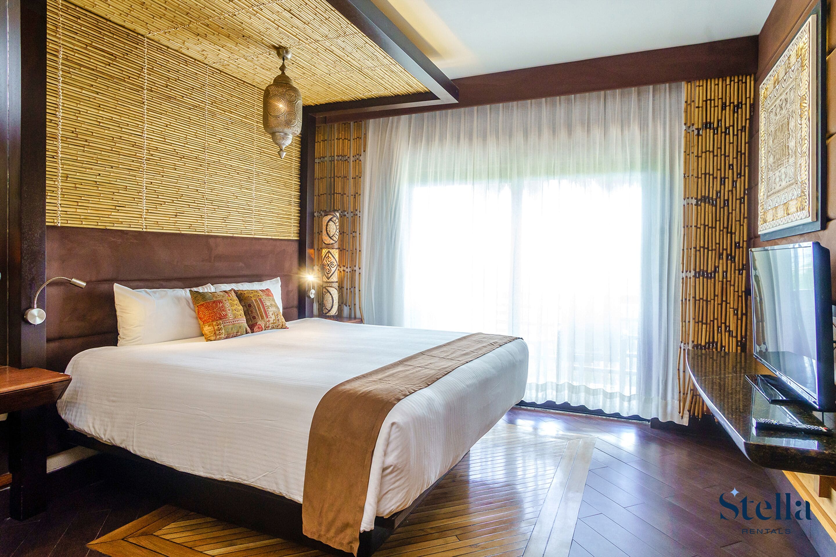 Una cama grande con cabecera de madera, una mesita de noche, un televisor y una ventana con cortinas translúcidas.
