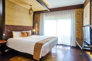 A large bed with a wooden headboard, a bedside table, a television, and a window with sheer curtains.