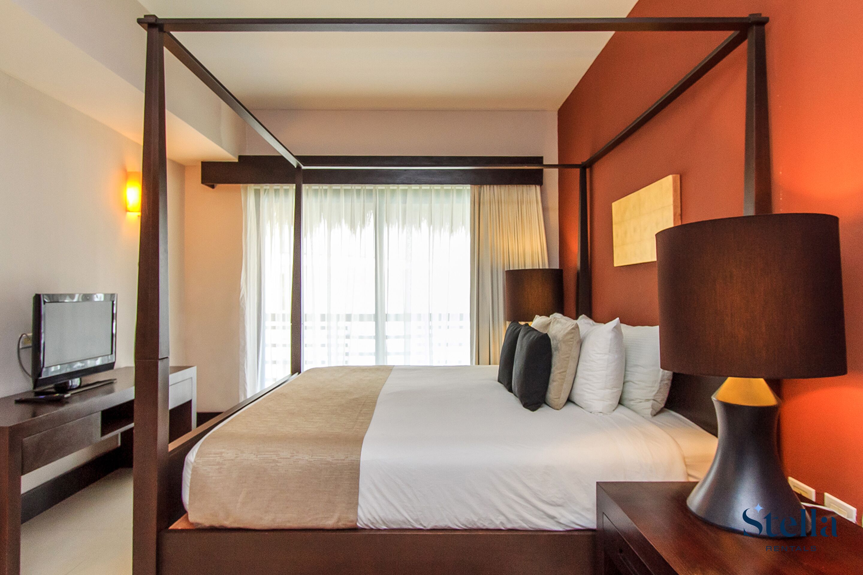 A four-poster bed with a television on a stand to the left, a bedside table with a lamp, and a window with curtains.