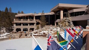 A large building with multiple balconies and a stone facade, surrounded by snow and trees. Flags are displayed in front of the building.
