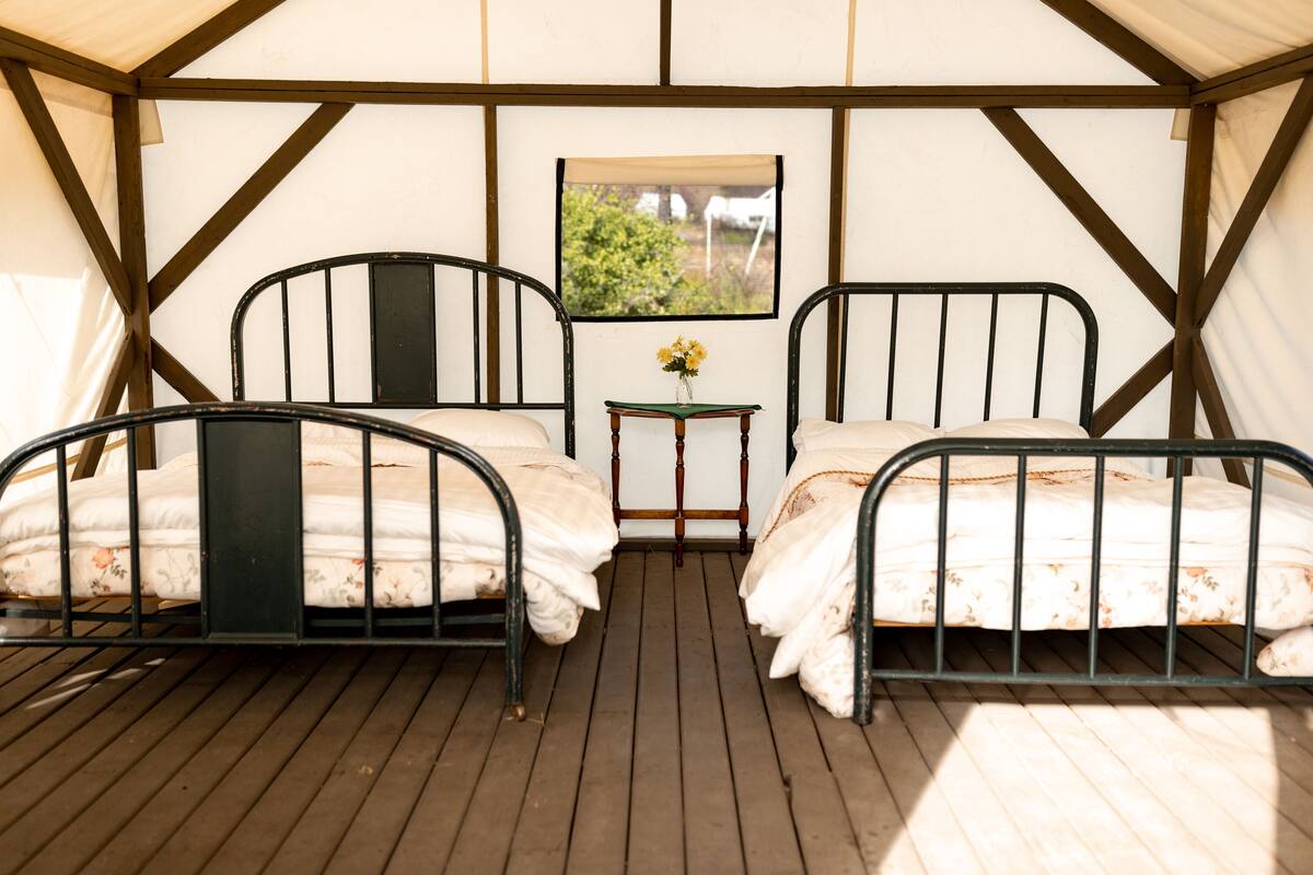 A tented accommodation with two metal-framed beds, a small wooden table with a vase of flowers, and a window with a view of greenery.