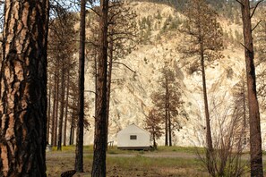 A small, single-story cabin with a gabled roof, surrounded by tall trees and a mountainous backdrop.