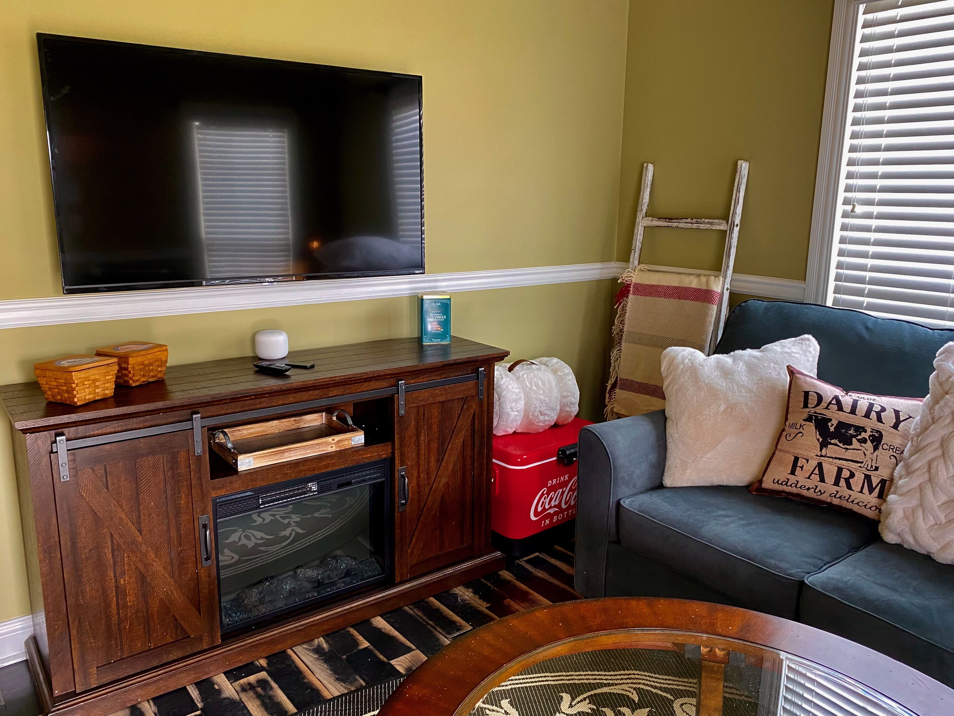 Una sala de estar con un televisor de pantalla plana montado en la pared, un mueble de madera con horno integrado, un sofá gris con almohadones decorativos y un refrigerador rojo Coca-Cola.