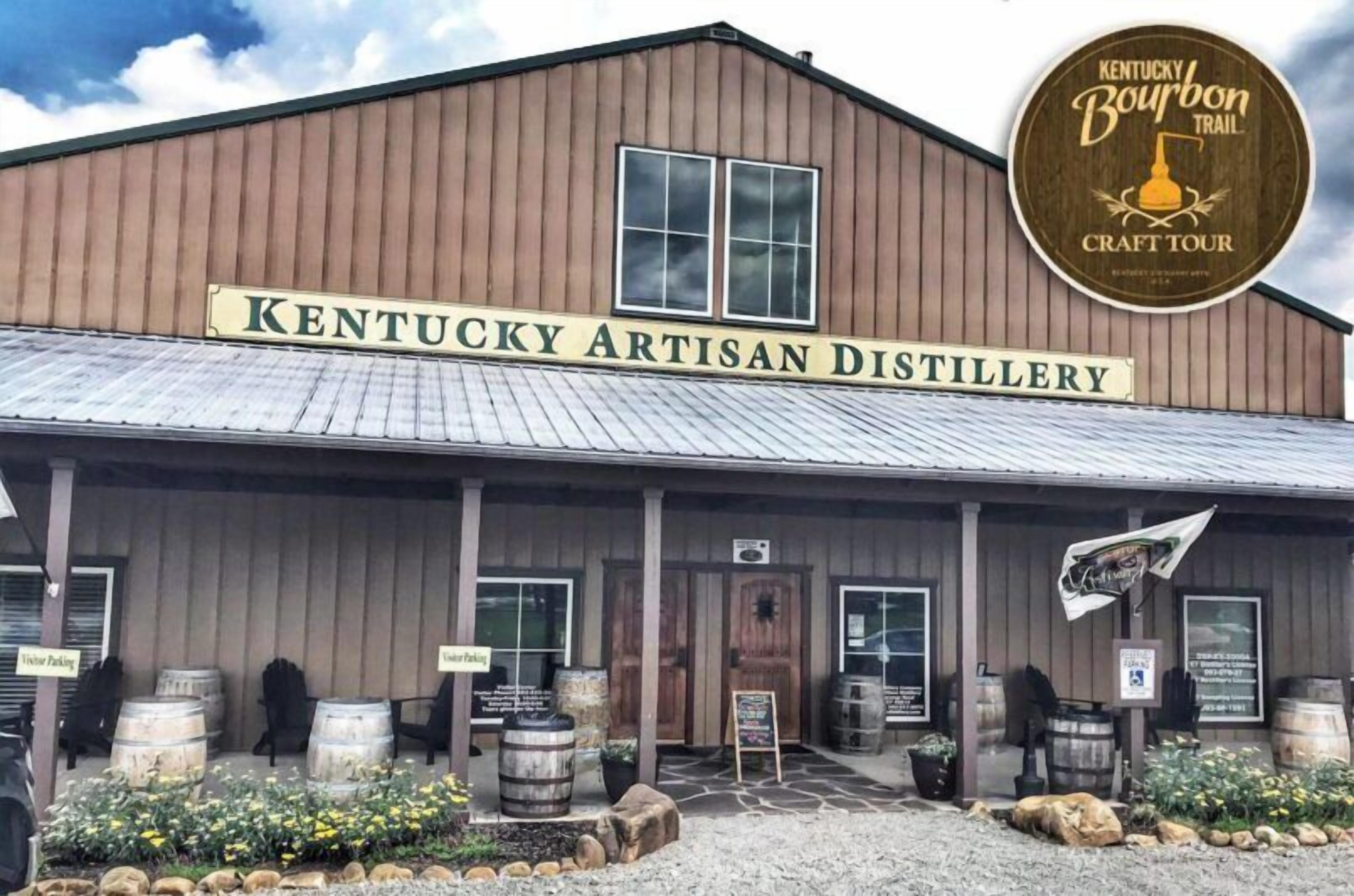 Het gebouw van Kentucky Artisan Distillery met een rustieke buitenkant, houten vaten en een bord voor bezoekersparkeren.
