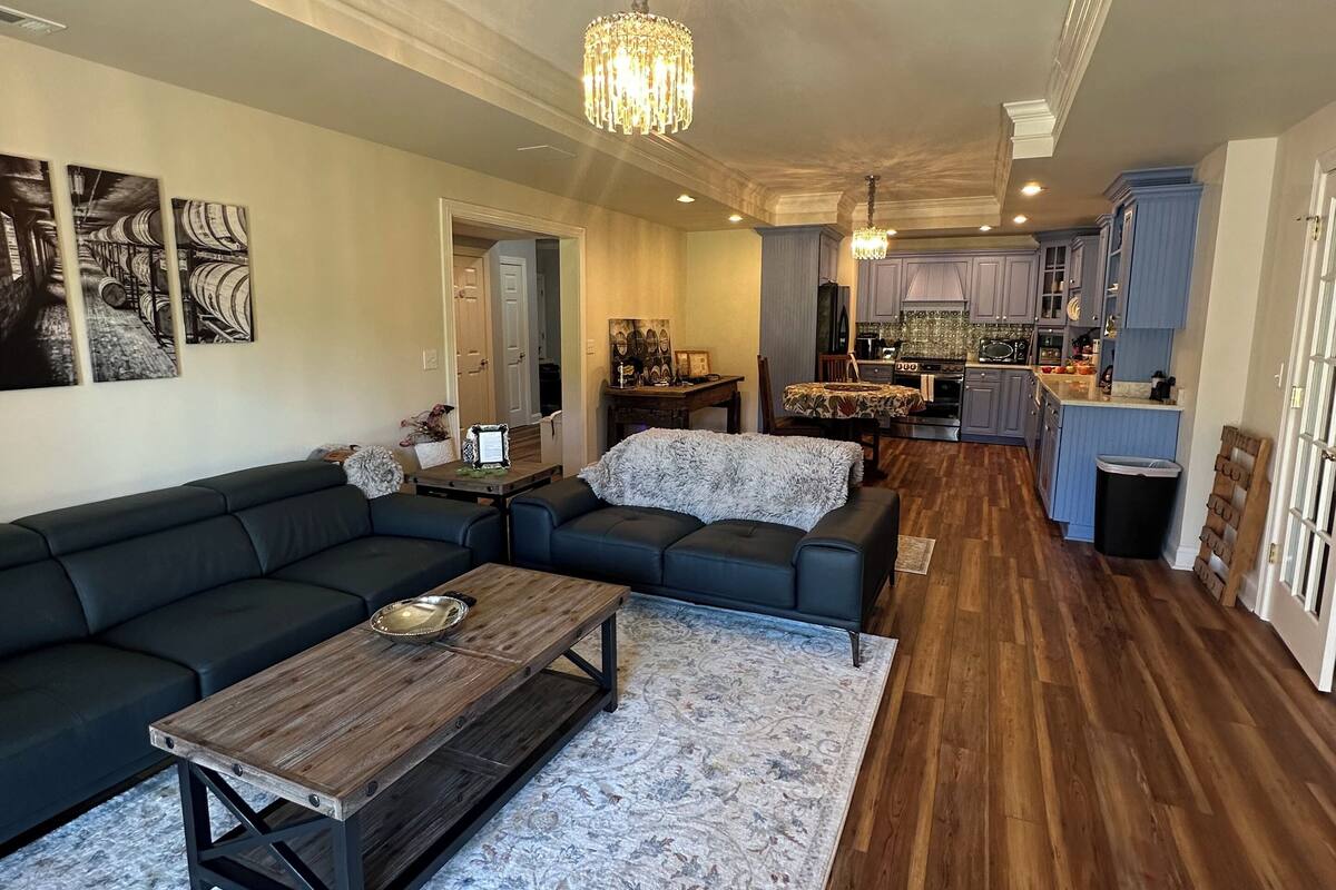 A spacious living room with a dark-colored sectional sofa, a wooden coffee table, and a patterned rug. A chandelier hangs from the ceiling, and the room features a wooden floor and a kitchen area with blue cabinets.