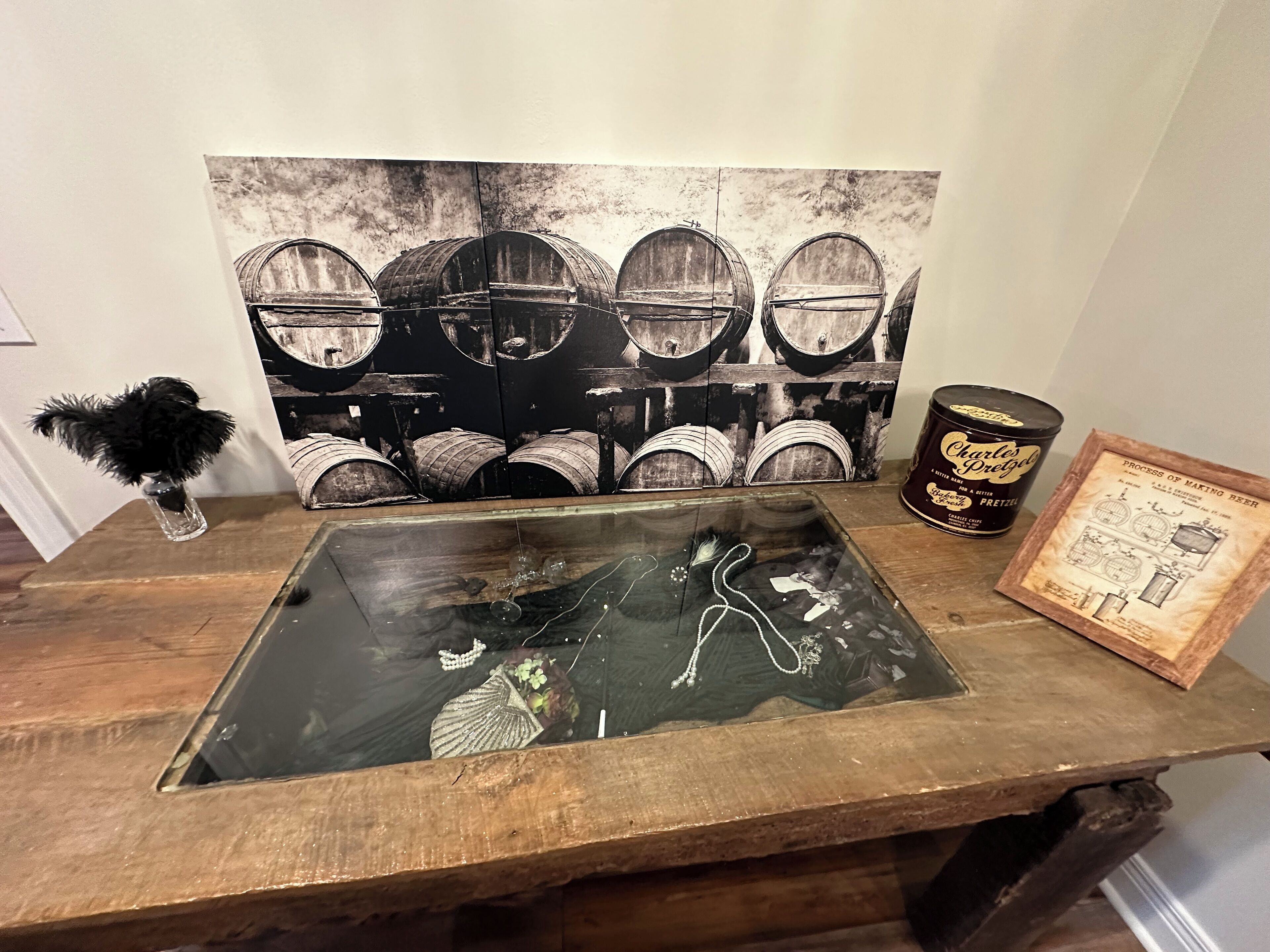 Una mesa de madera con cubierta de vidrio, una obra de arte blanco y negro de barriles, una lata de pretzels Charles & Evelyn y un documento de patente enmarcado.