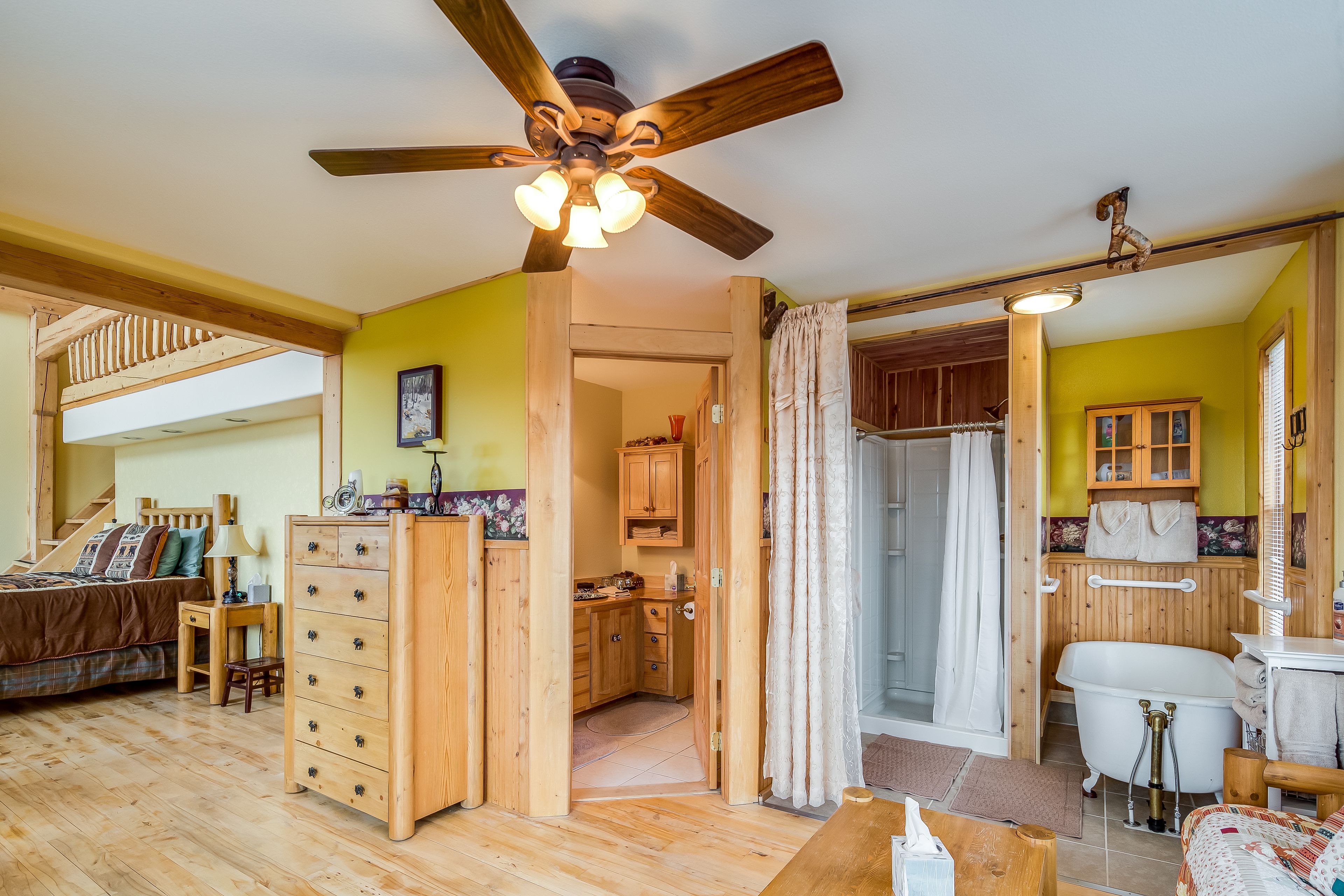 A cozy room with a wooden ceiling fan, a bed with a plaid comforter, a wooden dresser, and a small kitchen area with wooden cabinets and a white bathtub.