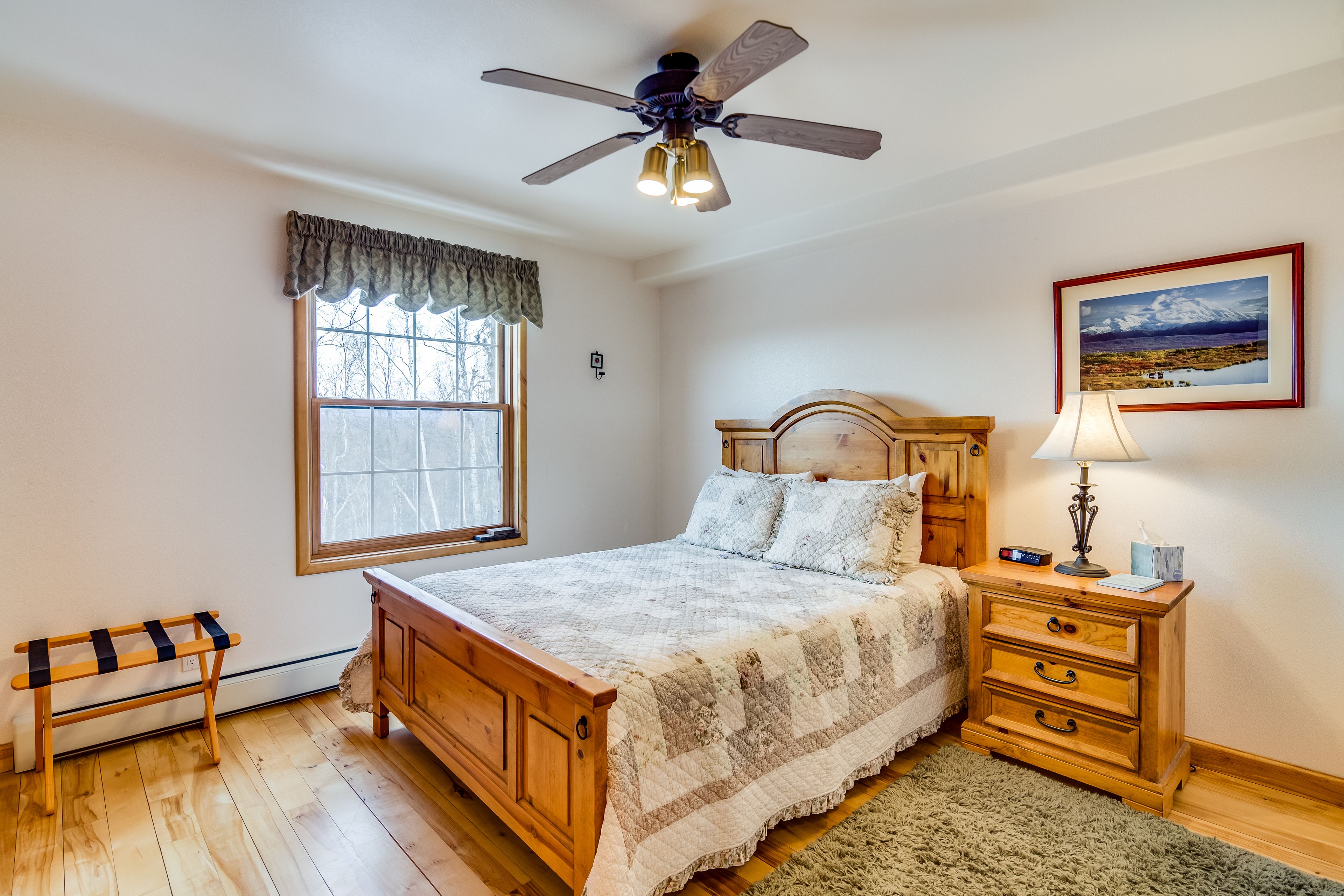 A bedroom with a wooden bed, a nightstand with a lamp, a window with a view, and a ceiling fan.