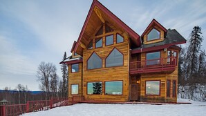 A large wooden cabin with multiple windows and a balcony, surrounded by snow.