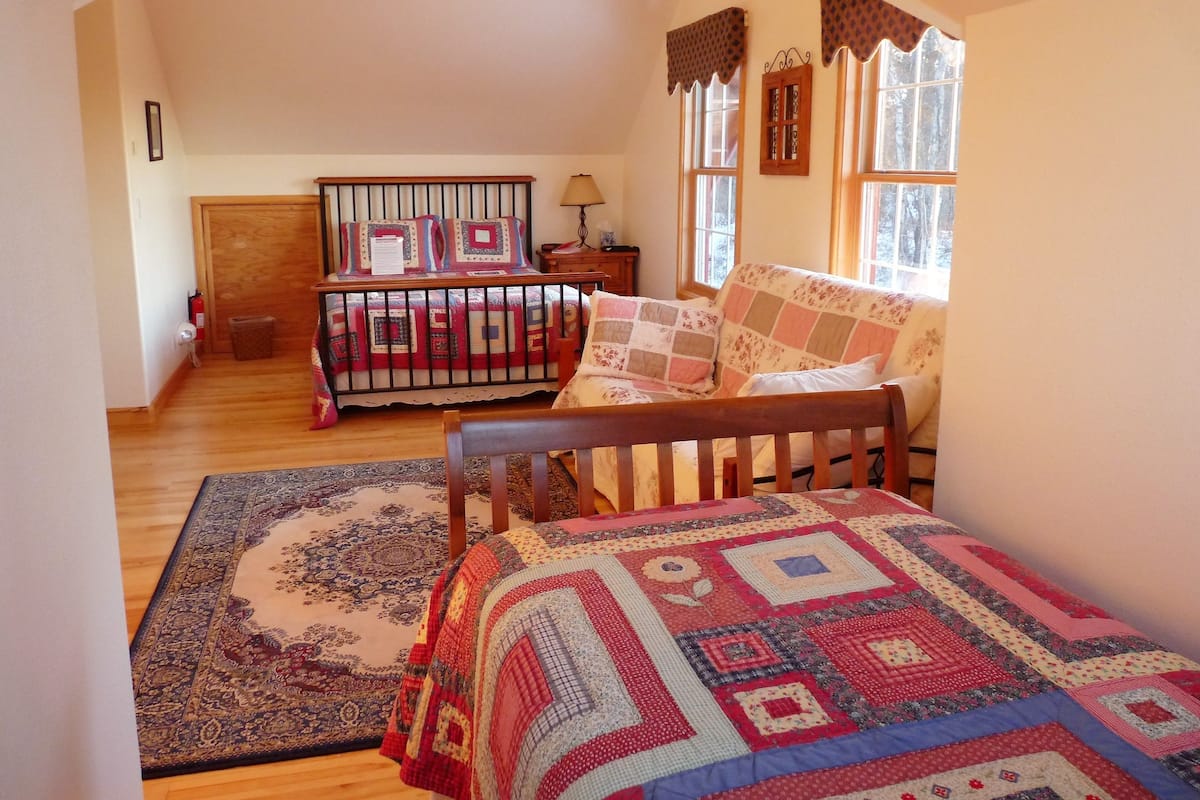 Quarto aconchegante com cama de madeira, um banquinho combinando, tapete estampado e janela com cortinas.